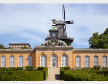 Historisches Gebäude mit gelb-orangefarbener Fassade, weißen hohen Fenstern und dekorativen Verzierungen, dahinter eine große Holz-Windmühle. Im Vordergrund führen geschnittene Hecken entlang eines Gehwegs.
