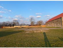 Weite Wiese mit vereinzelten gepflasterten Flächen, im Hintergrund eine Mauer, Bäume und mehrere flache Gebäude mit hellen Dächern. Rechts im Bild steht ein großes, langes Backsteingebäude mit rotem Satteldach. Der Himmel ist hellblau mit vereinzelten Wolken.