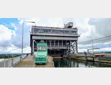 Großes historisches Schiffshebewerk aus grauem Metall mit verglasten Fenstern, im Vordergrund ein grünes, historisches Zugmaschinenfahrzeug auf einem Holzsteg. Der Ort ist von Wasserkanälen und einer weiten Landschaft umgeben, unter einem bewölkten Himmel.