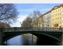 Eine grüne Metallbrücke mit verzierten Geländern überspannt einen Fluss, gesäumt von kahlen Bäumen und historischen Wohnhäusern mit gelben und weißen Fassaden. Straßenlaternen stehen entlang der Brücke, und einige Menschen sind auf ihr zu sehen.