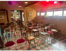 Ein kleiner Veranstaltungsraum mit gemustertem Fliesenboden, einer Mischung aus bunt gepolsterten Holzstühlen und einigen Pflanzen auf der Fensterbank. Die Fenster an der rechten Wand lassen Tageslicht herein, während an der Decke bunte Bühnenlichter sichtbar sind.