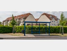 Bushaltestelle mit blauem Unterstand aus Glas und Metall, umgeben von einer hohen Hecke und jungen Bäumen. Im Hintergrund befinden sich mehrstöckige Wohngebäude mit roten Ziegeldächern und Balkonen.