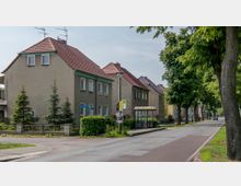 Straßenszene in einem Wohngebiet mit Reihenhäusern und rotem Ziegeldach, daneben eine Bushaltestelle mit Überdachung. Die Straße wird von großen Bäumen gesäumt, und ein Gehweg verläuft parallel zur Fahrbahn.