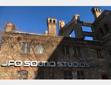 Backsteingebäude mit verwitterten Mauern und teilweise überwachsenen Fenstern, auf dessen Fassade der Schriftzug „UFO SOUND STUDIOS“ in silbernen Buchstaben angebracht ist; im Hintergrund ragen Ziegelkamine und Ruinenelemente in den blauen Himmel.