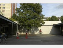 Ein kleiner öffentlicher Platz mit einem großen, grünen Baum in der Mitte, flankiert von einem flachen Gebäude mit Flachdach und weiß gekachelter Fassade. Im Hintergrund ist ein mehrstöckiges Wohngebäude zu sehen, während im Vordergrund ein Mann auf einem Fahrrad vorbeifährt und Coca-Cola-Stehtische vor einem Imbiss stehen.