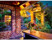 Gemütliche Terrasse mit roten Backsteinwänden und großen Glastüren, die in einen beleuchteten Innenraum führen. Im Vordergrund steht ein Holztresen mit einem Blumenstrauß, darüber eine geflochtene Hängelampe; im Hintergrund rankt Grünpflanzenbewuchs und Gartenmöbel sind sichtbar.