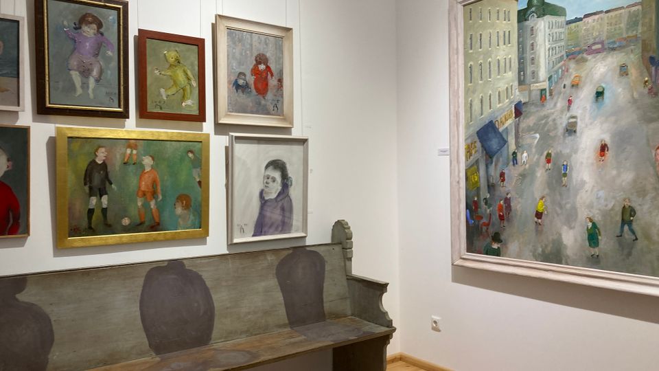 Ecke eines Kunstmuseums mit weißen Wänden und hellem Holzboden, an denen mehrere gerahmte Gemälde in Hängung zu sehen sind. Unter den Bildern steht eine lange, bemalte Holzbank, rechts hängt ein großes Gemälde mit einer Straßenszene zwischen hohen Gebäuden.