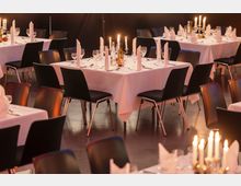 Ein festlich gedeckter Speisesaal mit mehreren Tischen, die weiß eingedeckt sind und mit Kerzenleuchtern, gefalteten Stoffservietten, Gläsern und Besteck dekoriert wurden. Die Tische sind von schwarzen Stühlen mit Metallbeinen umgeben, und die Beleuchtung ist warm und stimmungsvoll.
