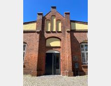 Backsteinfassade eines historischen Gebäudes mit Spitzbögen und gelben Putzfeldern. Ein moderner, schwarzer Eingang ist eingebaut; daneben befinden sich ein Schild und die Hausnummer 5.