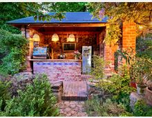 Kleiner offener Kiosk aus Backstein mit Holzdach, dekorativen Hängelampen und einer Theke mit Fliesenakzenten. Umgeben von grüner Bepflanzung und Bäumen in einer Gartenszene.