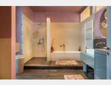 Modernes Badezimmer mit pastellfarbenen Wänden in Lila und beigen Fliesen. Das Zimmer umfasst eine bodenebene Glasdusche, eine Badewanne, einen Waschtisch mit ovalem Spiegel, einen Handtuchheizkörper und dunklen Fliesenboden mit Teppichakzenten.