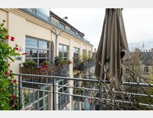 Mehrstöckiges Wohngebäude mit beigefarbener Fassade, großen Fensterfronten und Balkonen, die mit Pflanzen und Blumenkästen dekoriert sind. Im Vordergrund ein geschlossener Sonnenschirm auf einem Balkon sowie Blick auf umliegende Dächer und den Berliner Fernsehturm im Hintergrund.