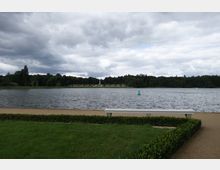 Ein See mit bewölktem Himmel, umgeben von Bäumen und einer Rasenfläche im Vordergrund. Eine gerade Uferlinie mit einer Bank und einer gepflegten Hecke grenzt an den See, in dem eine grüne Boje schwimmt.