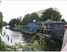 Eine Wasserstraße mit einem kleinen, blau-rot gestrichenen Gebäude am Ufer, das auf Stelzen über dem Wasser steht. Im Hintergrund sind Bäume, ein anlegendes Boot und ein größerer, heller Gebäudekomplex zu sehen.