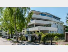 Moderne Wohnhausfassade mit mehreren Balkonen und großen Fenstern, umgeben von grüner Vegetation und Bäumen. Im Vordergrund sind ein Gartenzaun, Müllcontainer und ein Gehweg sichtbar.