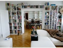 Wohnzimmer mit weißen Bücherregalen, die Bücher und CDs enthalten, einem hellen Parkettboden und einer weißen Couch mit einer Felldecke. Im Hintergrund ist ein Essbereich mit einem Holztisch und Stühlen sowie einer Glasvitrine zu sehen.