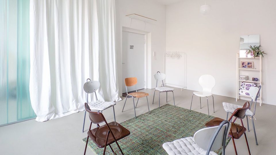 Heller, minimalistischer Innenraum mit grauem Boden und weißen Wänden, in dessen Mitte mehrere Stühle im Kreis um einen grünen Teppich stehen. Links hängt ein bodenlanger weißer Vorhang vor großen Fensterflächen, rechts sind eine Tür, eine Garderobe und ein Regal mit kleinen Gegenständen zu sehen.