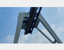 Industrieller Hafenkran aus Metall mit einer großen, offenen Konstruktion und schräg verlaufenden Stützpfeilern vor einem teilweise bewölkten blauen Himmel.