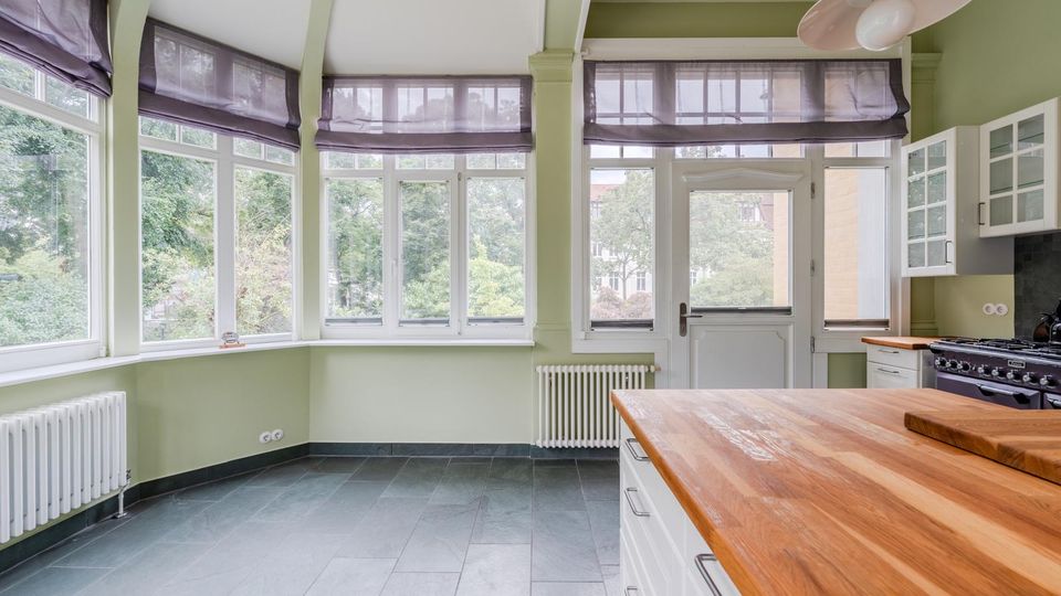 Heller Küchenraum mit großen, umlaufenden Fenstern und halbtransparenten Raffrollos, hellgrünen Wänden und grauem Fliesenboden. Rechts sind weiße Küchenschränke, ein Herd sowie eine breite Holzarbeitsplatte zu sehen, daneben eine Glastür nach außen.