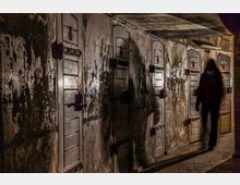 Dunkler, gewölbter Gang mit einer Reihe kleiner, hölzerner Türen mit Metallbeschlägen an einer abgenutzten Betonwand. Eine verschwommene menschliche Silhouette ist im Halbdunkel zu sehen.