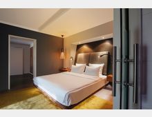 Modernes Hotelzimmer mit einem großen Doppelbett, dunkelgrauer Wand, gepolstertem Kopfteil mit integrierter Beleuchtung und einer hängenden Lampe. Auf dem Boden liegt ein gelber Teppich, und eine offene Tür führt in einen angrenzenden Raum.