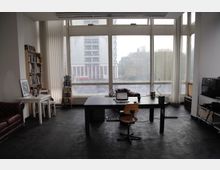 Ein helles Arbeitszimmer mit großen bodentiefen Fenstern, die auf eine städtische Umgebung blicken. Der Raum enthält einen schwarzen Schreibtisch mit einem Stuhl, ein Regal mit Büchern, ein kleines weißes Tischset sowie eine braune Chesterfield-Couch.