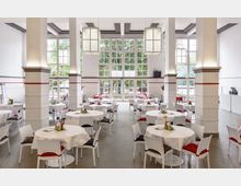 Großer, heller Saal mit hohen Decken, weißen Wänden und großen Fenstern, die Blick auf grüne Bäume ermöglichen. Der Raum ist mit runden Tischen, weißen Stühlen und schlichten Dekorationen ausgestattet, die Tische sind mit weißen Tischdecken bedeckt.