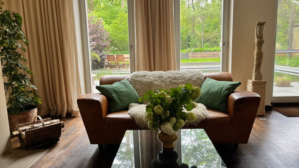 Helles Wohnzimmer mit bodentiefen Fenstern und beigen Vorhängen, davor ein braunes Ledersofa mit grünen Kissen und einer weißen Decke sowie ein Glastisch mit Blumen. Durch die Fenster ist eine Terrasse und ein grüner Garten mit Bäumen zu sehen; daneben stehen eine Zimmerpflanze, ein Korb mit Holzscheiten und eine Skulptur auf einem Sockel.