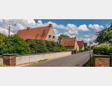 Straßenszene in einem Wohngebiet mit Einfamilienhäusern im traditionellen Baustil, erkennbar an ihren roten Satteldächern und hellen Fassaden. Die Straße ist asphaltiert, von Hecken und kleinen Mauern eingefasst, und der Himmel ist leicht bewölkt.