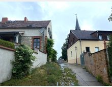 Gepflasterte, leicht ansteigende Gasse in einem Dorf mit zwei Gebäuden am Rand: links ein Haus mit Natursteinfassade und roten Ziegelakzenten, rechts eine helle Fassade mit Holzverkleidung im oberen Bereich. Im Hintergrund ein Kirchturm mit Wetterhahn sowie eine Straßenlaterne.
