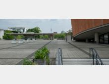 Weitläufiger, gepflasterter Platz mit modernen architektonischen Elementen, darunter ein rundlich geschwungenes Gebäude mit orangefarbener Fassade rechts und eine Überdachung mit Säulen in der Mitte. Im Hintergrund sind Bäume, der Berliner Fernsehturm und die Glaskuppel des Reichstagsgebäudes erkennbar.