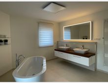 Modernes Badezimmer mit freistehender Badewanne, Doppelwaschbecken auf einer Holzplatte und beleuchtetem Wandspiegel. Das Zimmer hat helle Wände, eine mattierte Fenstergardine und einen Handtuchheizkörper rechts.