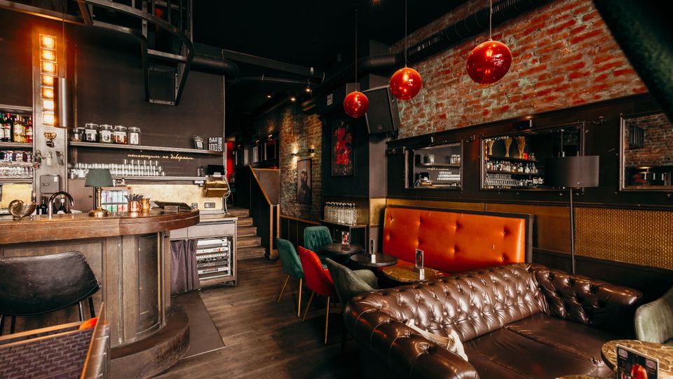 Innenansicht einer Bar im Industrial-Stil mit Backsteinwand, dunkler Decke und schwarzer Metall-Spindeltreppe. Zu sehen sind eine Holztheke mit Flaschenregalen, rote Kugel-Pendelleuchten sowie Lounge-Sitzplätze mit Chesterfield-Ledersofa und kleinen Tischen.