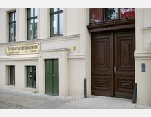 Fassade eines historischen Gebäudes mit beigem Putz und grün gestrichenen Fenstern und Türen. Rechts im Bild befindet sich ein großes, verziertes Holztor, links ein Schild mit der Aufschrift „Antiquariat Alt-Köpenick“.