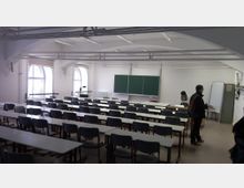 Ein heller Seminarraum mit Reihen aus grauen Tischen und Stühlen, großen Rundbogenfenstern an der linken Wand, einer grünen Tafel an der Vorderseite und einem Overheadprojektor. Zwei Personen sind im Raum zu sehen, eine steht und eine arbeitet an einem Tisch.