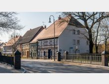 Straßenszene mit historischen Fachwerkhäusern und modernen Putzfassaden, daneben eine Fußgängerbrücke mit Metallgeländer und Backsteinpfeilern; im Hintergrund sind Bäume ohne Blätter sichtbar.