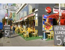 Straßenszene mit einem indischen Restaurant, das gelbe Stühle und Tische auf einer Grünfläche vor der Fassade aufgestellt hat. Ein großes Schild weist auf ein Mittagsangebot hin, während über dem Eingang ein Schild mit der Aufschrift „Indische Spezialitäten Küche“ hängt; daneben ein farbenfrohes Wandbild von Ganesha.