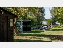 Uferbereich mit Bootsanlegern und kleinen Booten, die entlang eines schmalen Kanals liegen, umgeben von Bäumen und Wiesen. Im Vordergrund ein Holzgebäude und ein überdachter Unterstand, im Hintergrund ein See sichtbar.