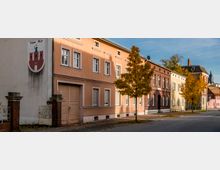 Straßenansicht mit einer Häuserreihe aus Altbauten in verschiedenen Pastellfarben und roten Backsteinen. Vor den Gebäuden stehen Bäume mit Herbstlaub, und an einer Hauswand ist ein Wappen mit einem Turm und einer Figur zu sehen.