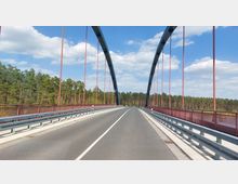 Straßenbrücke mit hoher Stahlbogenkonstruktion und roten Geländern, die über einen bewaldeten Bereich führt; blauer Himmel mit vereinzelten Wolken im Hintergrund.