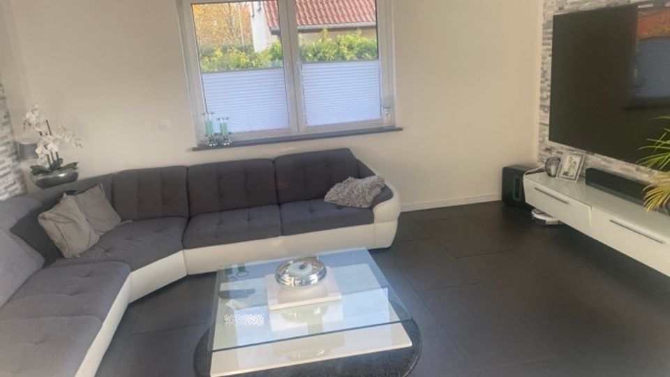 Modernes Wohnzimmer mit großem L-förmigem grauen Ecksofa, Glas-Couchtisch auf rundem Teppich und dunklem Fliesenboden. An der Wand befinden sich ein Fenster mit Plissees sowie ein wandmontierter Fernseher über einem weißen Lowboard vor einer grauen Steinoptik-Akzentwand.