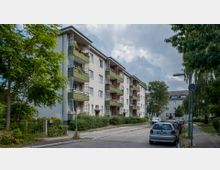 Wohnstraße mit mehrstöckigen Wohngebäuden im Hintergrund. Die Gebäude haben helle Fassaden, grüne Balkone und sind von Bäumen und Sträuchern umgeben; parkende Autos säumen die Straße.