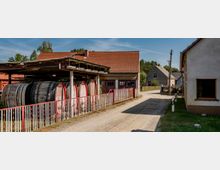 Eine ländliche Gasse mit Gebäuden im traditionellen Stil, rotem Ziegeldach und einem unter einem offenen Unterstand gelagerten Holzfässern. Im Hintergrund befinden sich weitere Häuser und Bäume, die die Siedlung umgeben.