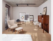 Minimalist bedroom or sitting area with a concrete ceiling, white walls, and woven carpet, lit by a window with dark blinds. Furnishings include a cream lounge chair with ottoman, two white pedestal chairs, a wood cabinet, and several framed artworks leaning against the back wall.