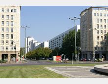 Stadtlandschaft mit breiter Straße, flankiert von mehrstöckigen Wohngebäuden mit heller Fassade. Im Hintergrund sind modernere Hochhäuser zu sehen, während im Vordergrund ein parkartiger Bereich mit Rasenflächen sichtbar ist.