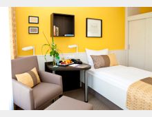 Helles, modernes Hotelzimmer mit einer gelb gestrichenen Wand, einem Einzelbett, einem kleinen runden Tisch, einer beigefarbenen Sitzgelegenheit sowie dekorativen Kissen und Gemälden. Auf dem Tisch stehen Blumen, Obst und eine Wasserflasche.