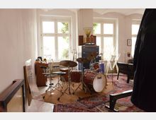 Ein heller Raum mit großen Fenstern und weiß gestrichenen Wänden, eingerichtet als Musikproberaum. Im Mittelpunkt steht ein Schlagzeug, umgeben von anderen Musikinstrumenten wie Trompeten und einer Klarinette. Der Boden ist mit bunten Teppichen bedeckt, und im Hintergrund sind Möbel wie ein Schrank und ein Verstärker sichtbar.