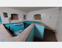 Ein leerer, gekachelter Raum mit einer halbkreisförmigen, türkisfarbenen Einfassung, die wie ein Pool oder Tauchbecken wirkt. Die Wände sind mit weißen Fliesen verkleidet, und es gibt mehrere kleine, bogenförmige Fensteröffnungen sowie einzelne angebrachte Wanddekorationen.