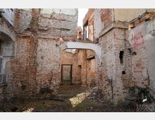 Innenhof einer verfallenen Backsteinruine mit freiliegenden, teils verputzten Mauern und einem gemauerten Rundbogen, der in einen weiteren Raum führt. Der Boden ist mit Moos und Schutt bedeckt, und mehrere Tür- und Fensteröffnungen sind ohne Rahmen oder Glas erhalten.