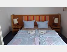 Doppelbett in einem schlichten Schlafzimmer mit buntem, gestreiftem Kopfteil, zwei blauen Kissen und farblich passenden Bettdecken. Auf den Bettdecken liegen gefaltete Handtücher mit kleinen Dekorationen, daneben zwei Nachttische mit Lampen.